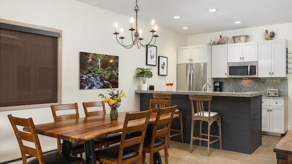 A modern open kitchen and dining area with a wooden table for six, a chandelier, white cabinetry, stainless steel appliances, and a gray island.