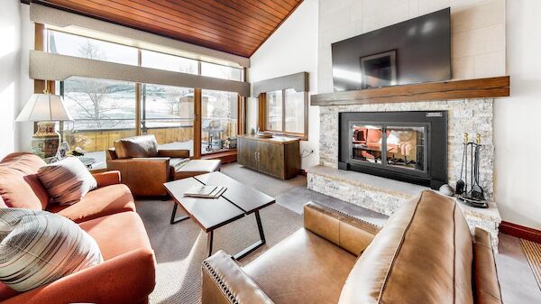 A cozy living room features a stone fireplace, large TV, and plush seating with expansive windows offering a scenic view.