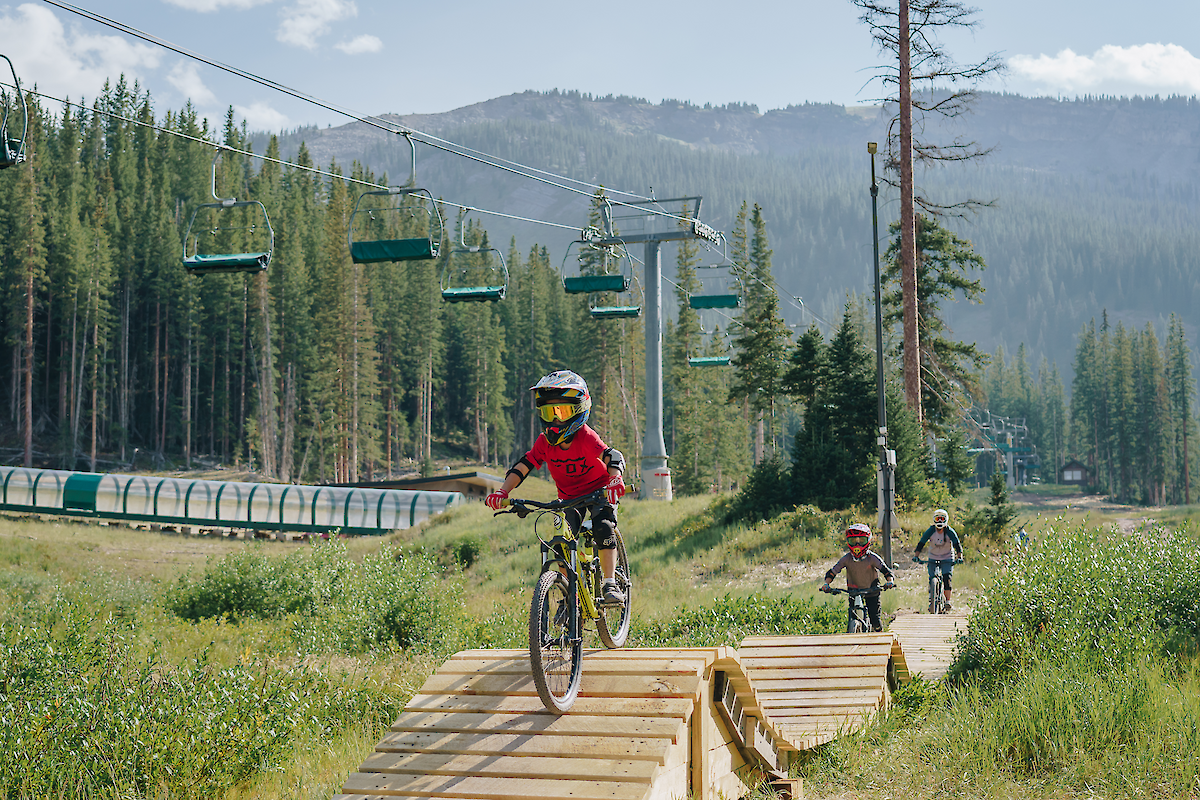 The image shows children biking on a wooden trail in a forested area, with ski lifts and mountains in the background.