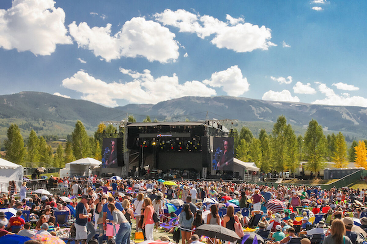 The image shows a large crowd at a music festival with a stage set against a scenic mountainous backdrop and clear skies.