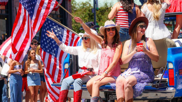 People at a parade waving American flags, wearing hats and boots, sitting on a float, enjoying the patriotic celebration.