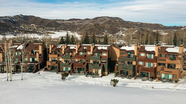 The image shows townhouses surrounded by snow, set against a backdrop of mountains and clear skies. It captures a serene winter landscape.