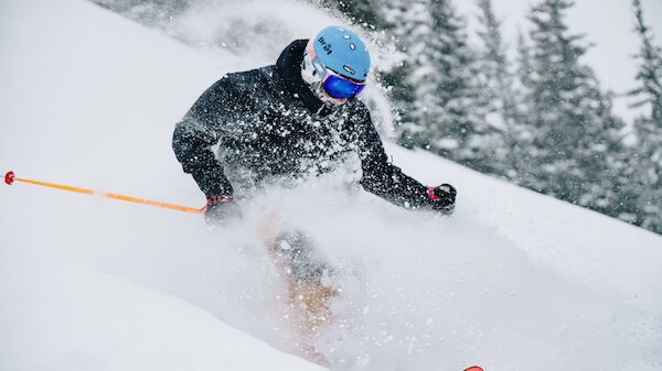A skier in a black jacket and blue helmet carving through fresh powder, kicking up snow as they race downhill with orange poles in a snowy forest.