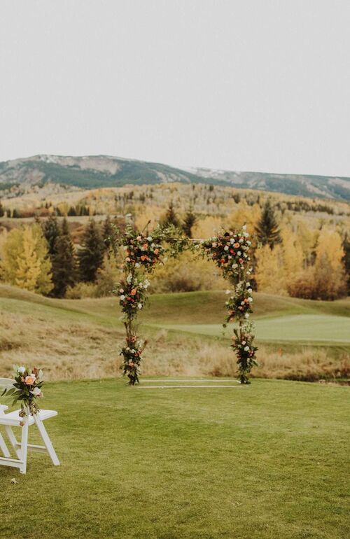 An outdoor wedding setup on a grassy field with white chairs lining a small aisle, floral arches, and a scenic autumn landscape backdrop.