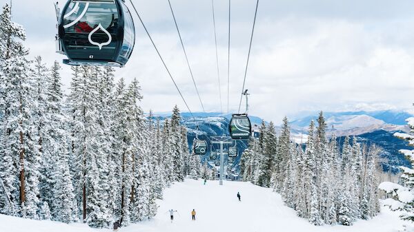 Snowy mountain slopes with frosted trees, a gondola lift gliding above, skiers below, and distant blue peaks under a cloudy sky.