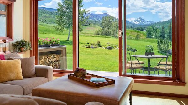 Cozy living room opening to a lush green lawn with mountains, a wood coffee table, couch with cushions, and a view through large glass doors.