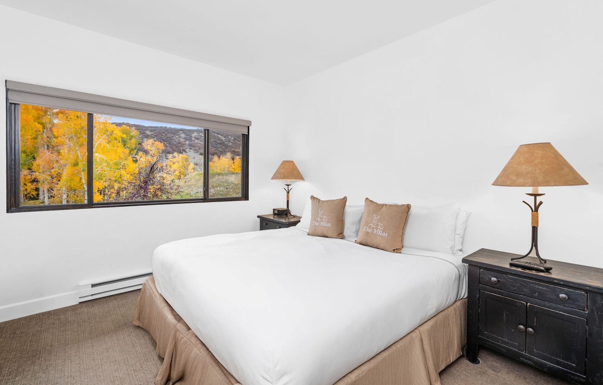 A bright, neatly made hotel-style bedroom with a white bed, brown headboard, two matching nightstands, lamps, and a window showing autumn trees outside.