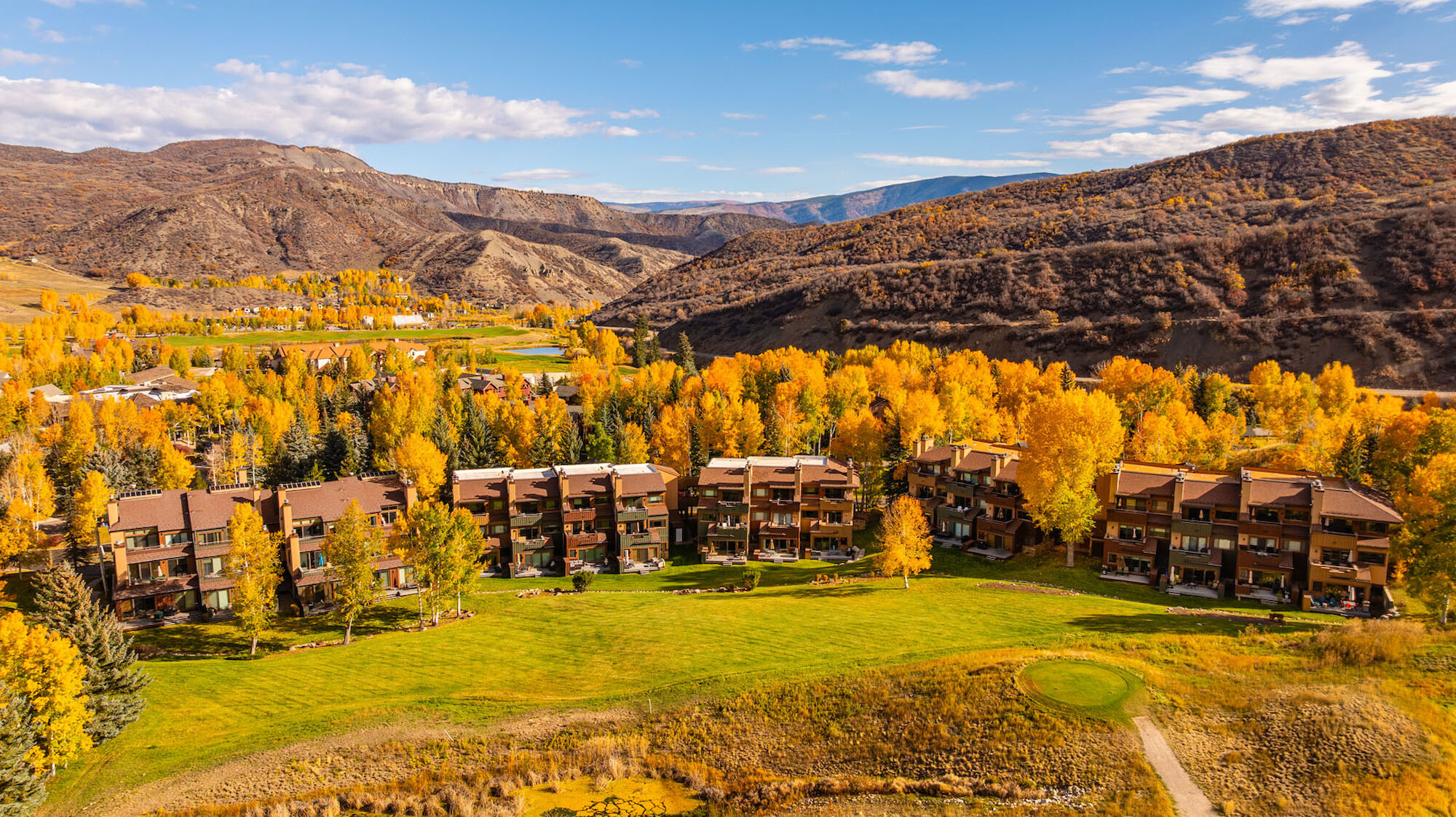 Autumn resort town with brown buildings surrounding a green field, golden aspen trees, and mountains in the background under a blue sky.