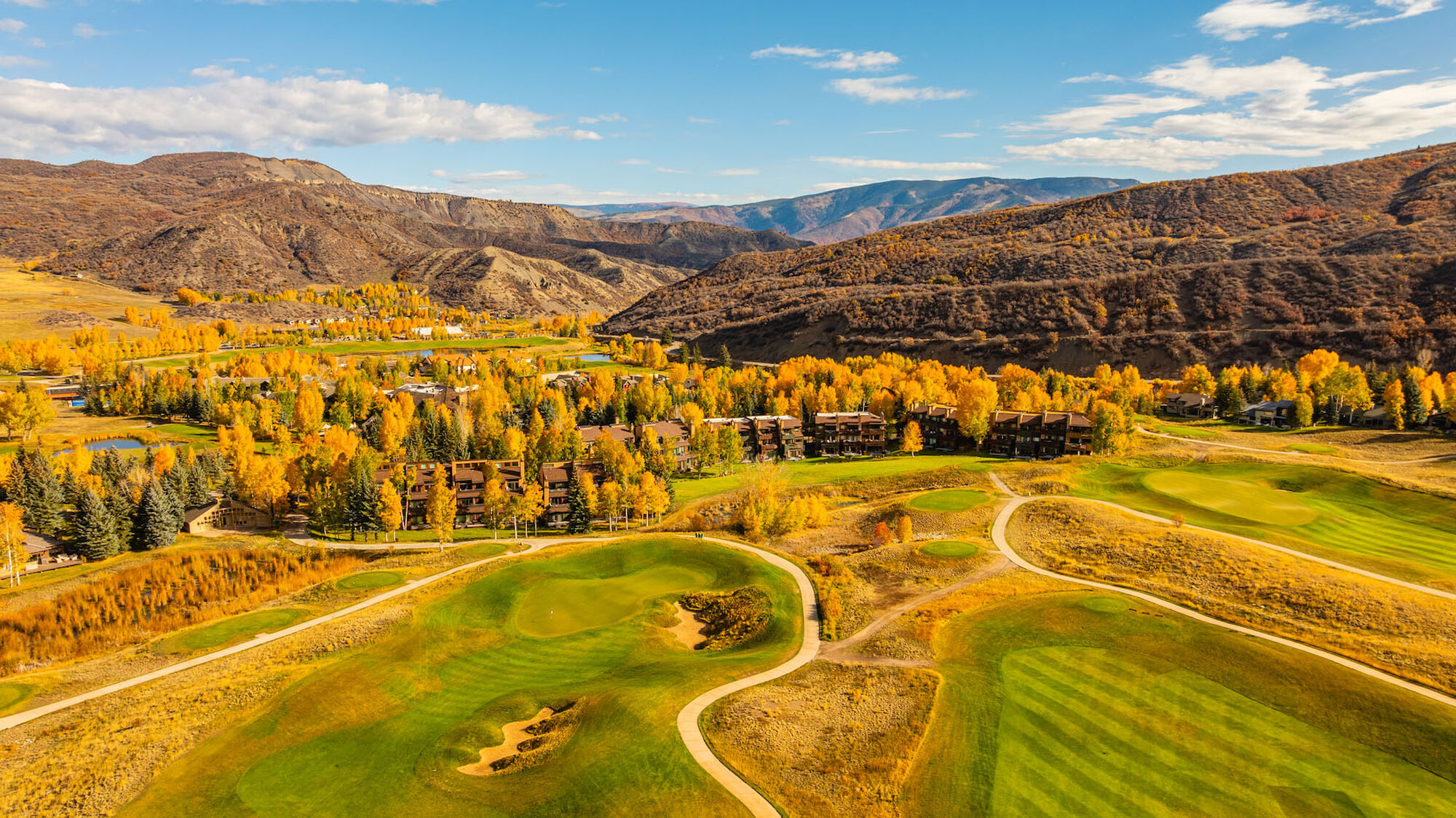 A sweeping desert valley golf course with winding fairways, autumn-yellow trees, and distant brown mountains under a blue sky.