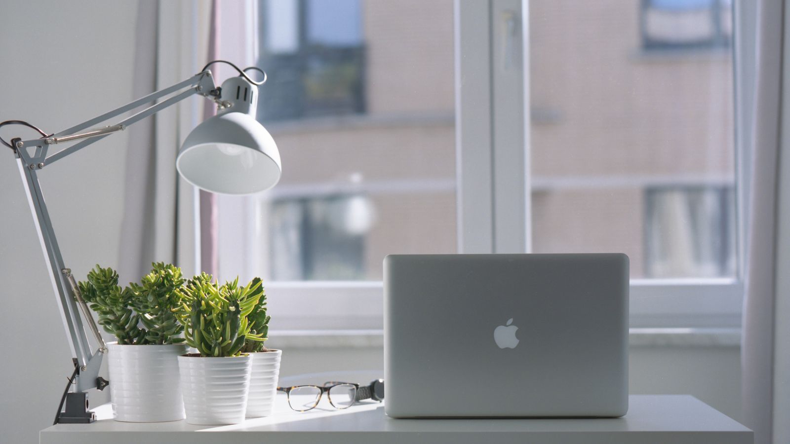 A minimalist workspace featuring a laptop, desk lamp, potted plants, and eyeglasses on a white desk by a window with curtains.