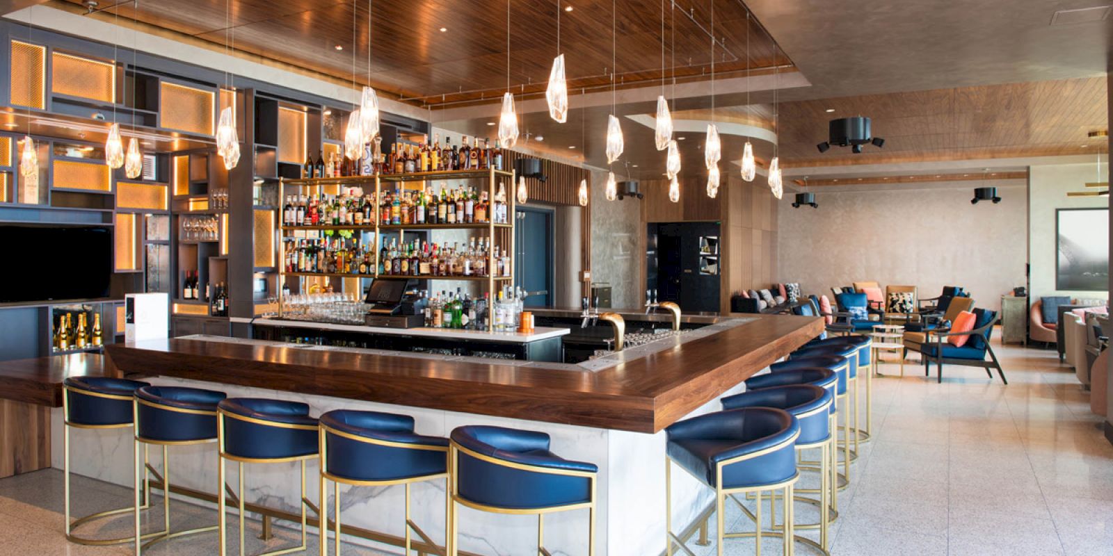 The image shows a modern bar with a large counter surrounded by blue and gold chairs, with shelves full of bottles and drop lights hanging.