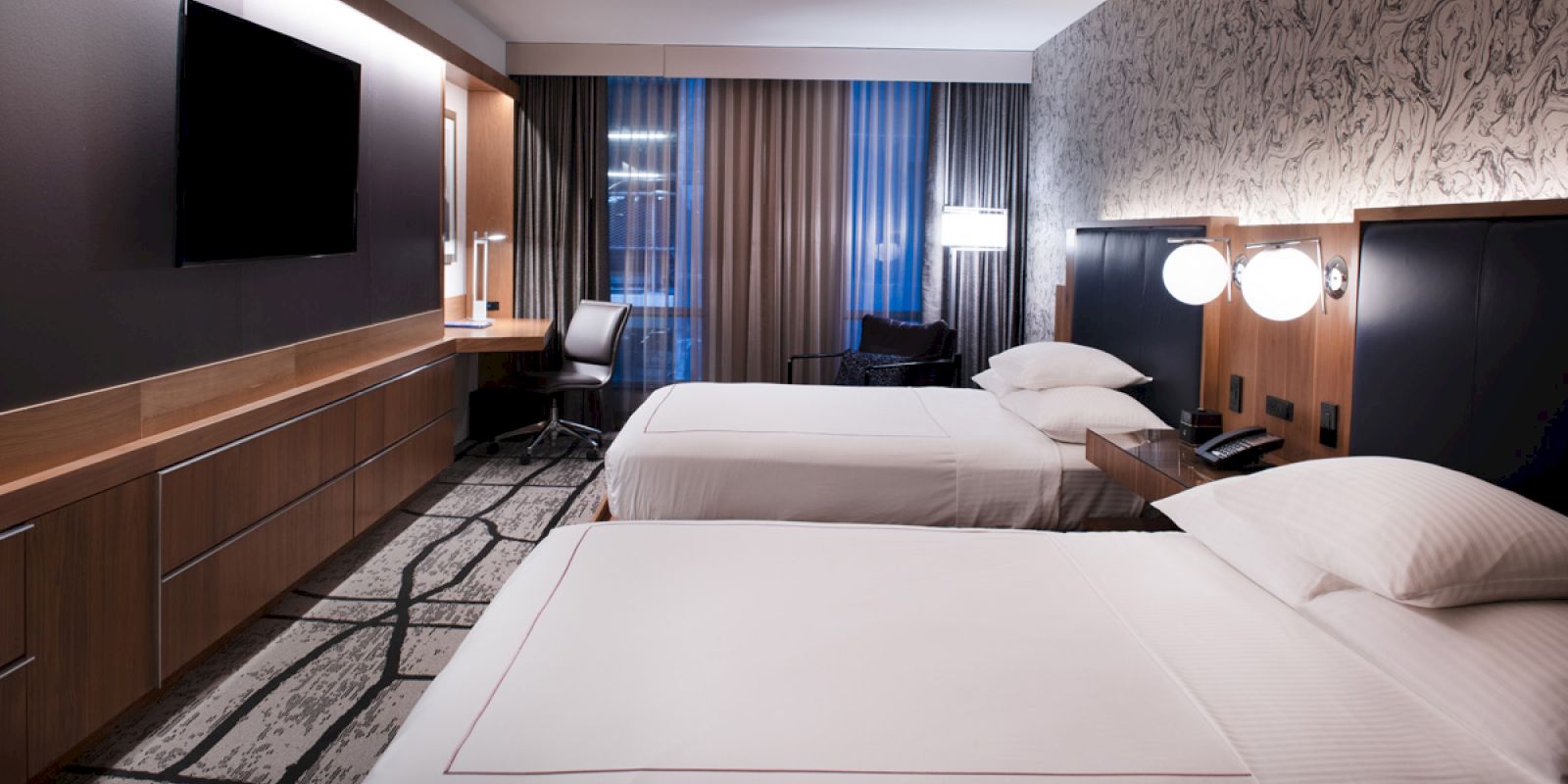The image shows a modern hotel room with two beds, a TV on the wall, a desk and chair, and a large window with curtains.
