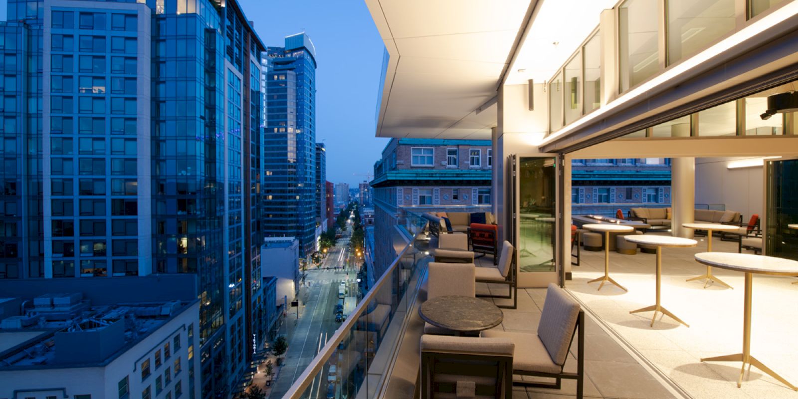 Cityscape view from a modern balcony with tables and chairs, looking down a street flanked by tall buildings during twilight.