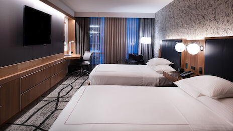 A modern hotel room with two beds, flat-screen TV, work desk, and stylish lighting. Large window with curtains, patterned walls, and cozy ambiance.