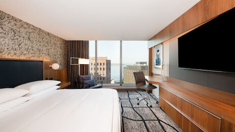 A hotel room with a large bed, modern desk area, flat-screen TV, and a view through a window showing buildings and part of a body of water.