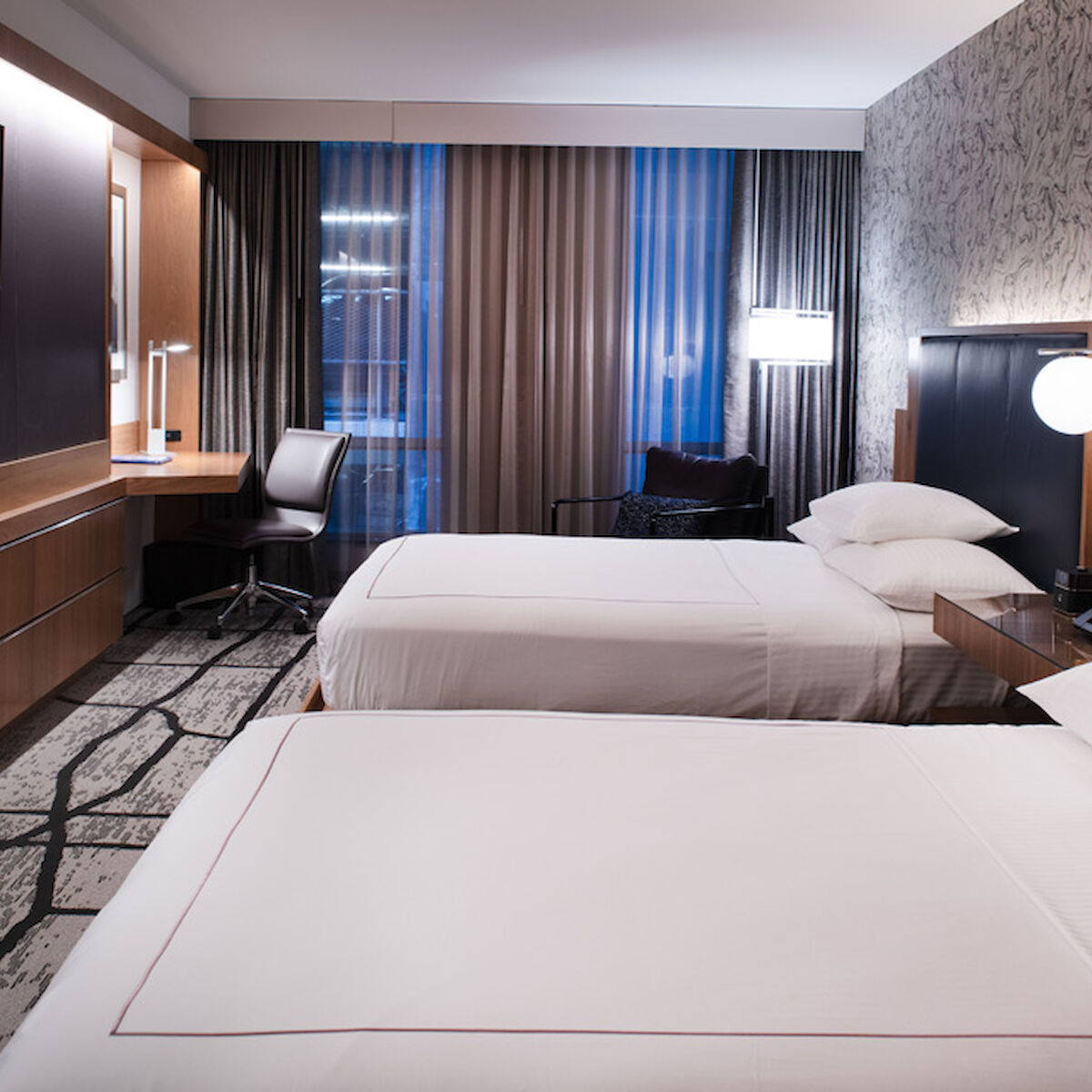 The image shows a hotel room with two beds, a wall-mounted TV, a desk, a chair, bedside lamps, and patterned wallpaper.