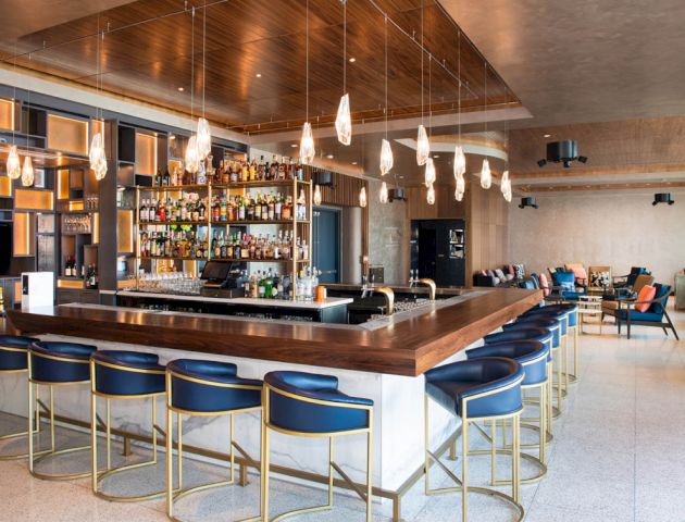This image shows a modern bar with wooden accents, blue bar stools, hanging lights, and a well-stocked shelf of liquor bottles in the background.