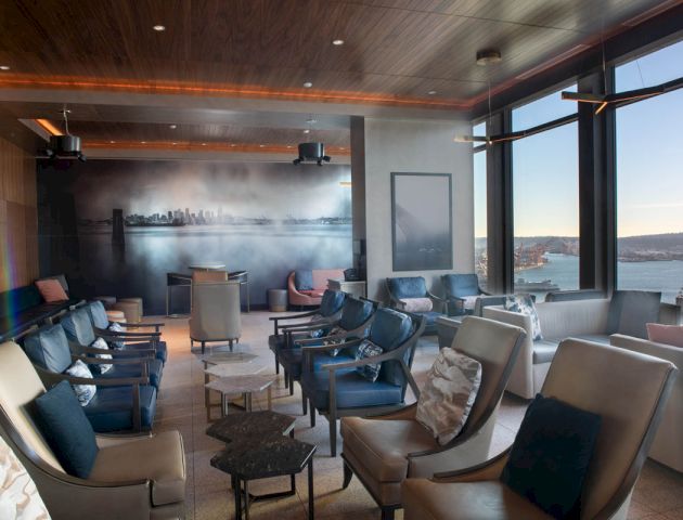 The image shows a luxurious lounge with ample seating, modern decor, large windows offering panoramic views, and a mural depicting a city skyline.