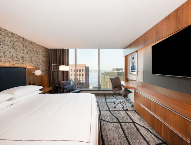 A modern hotel room with a large bed, wall-mounted TV, desk and chair, floor-to-ceiling windows, and a view of city buildings and the sea in the distance.