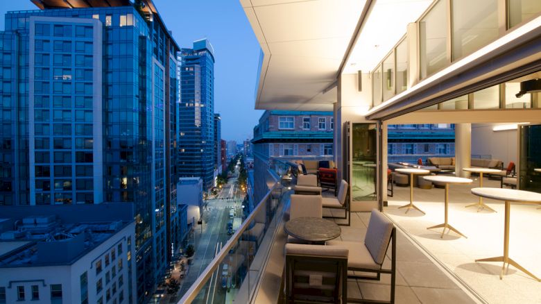 A modern cityscape at dusk with a high-rise building balcony featuring furniture and overlooking well-lit buildings and streets in the distance.