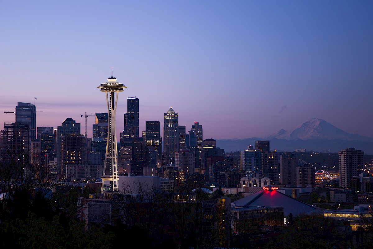 This is a cityscape showing the Seattle skyline at dusk, featuring the Space Needle and skyscrapers, with Mount Rainier visible in the background.