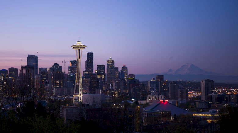 This is a cityscape showing the Seattle skyline at dusk, featuring the Space Needle and skyscrapers, with Mount Rainier visible in the background.