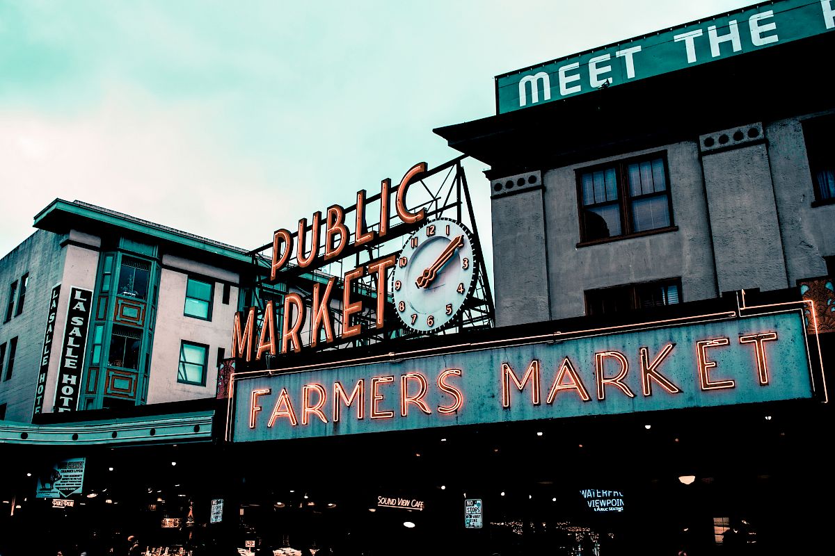 The image shows a neon sign for a Public Market and Farmers Market, with buildings in the background, suggesting an urban outdoor market setting.