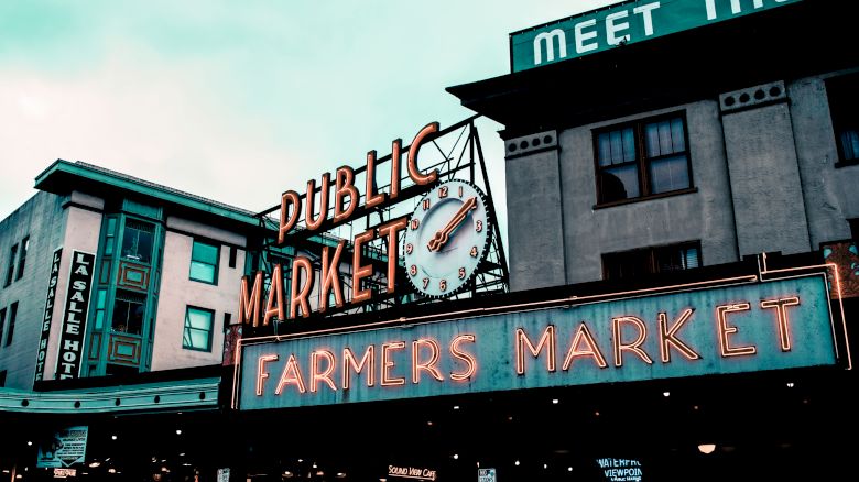 The image shows a neon sign for a Public Market and Farmers Market, with buildings in the background, suggesting an urban outdoor market setting.