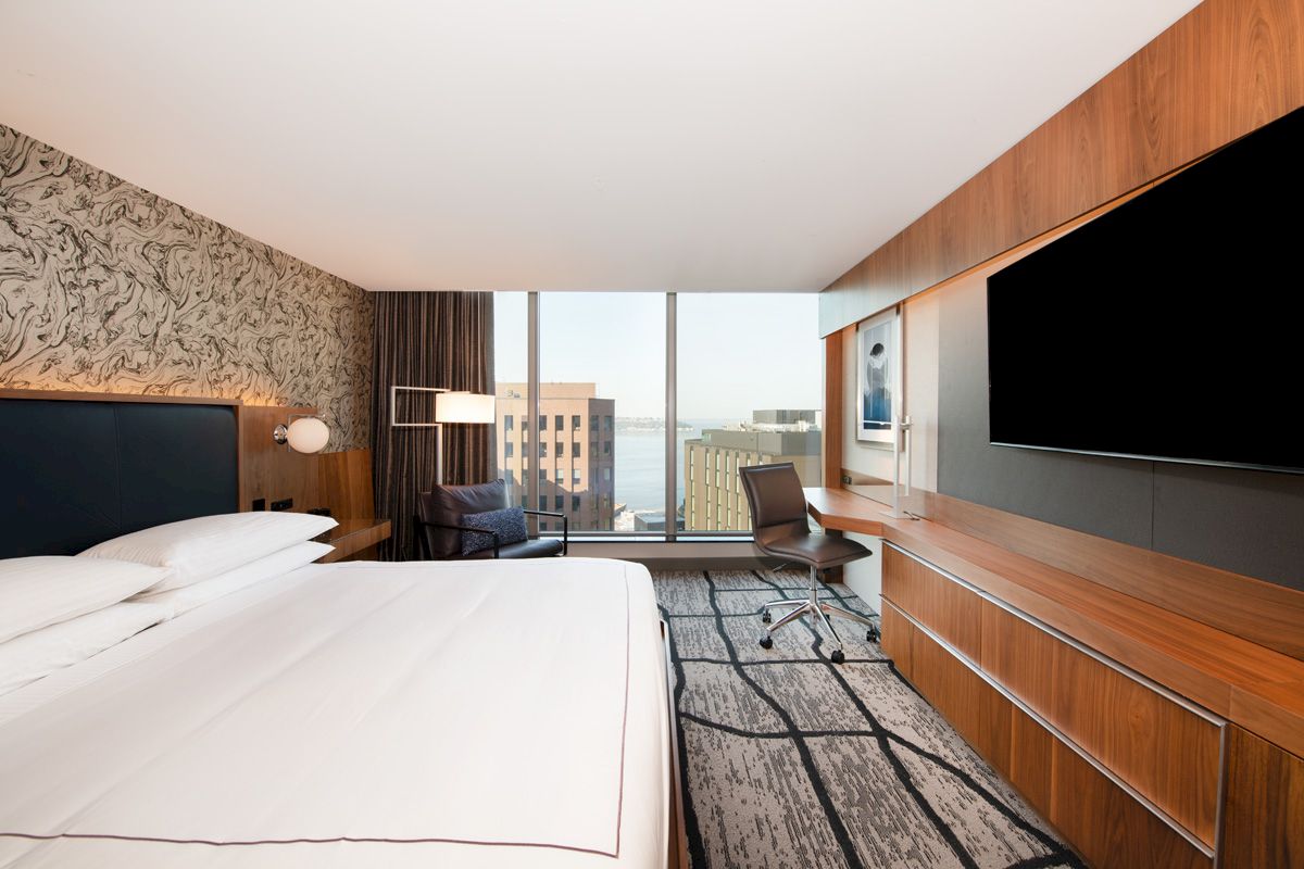 The image shows a modern hotel room with a large bed, a TV, a desk with a chair, a patterned carpet, and a window with a city view.