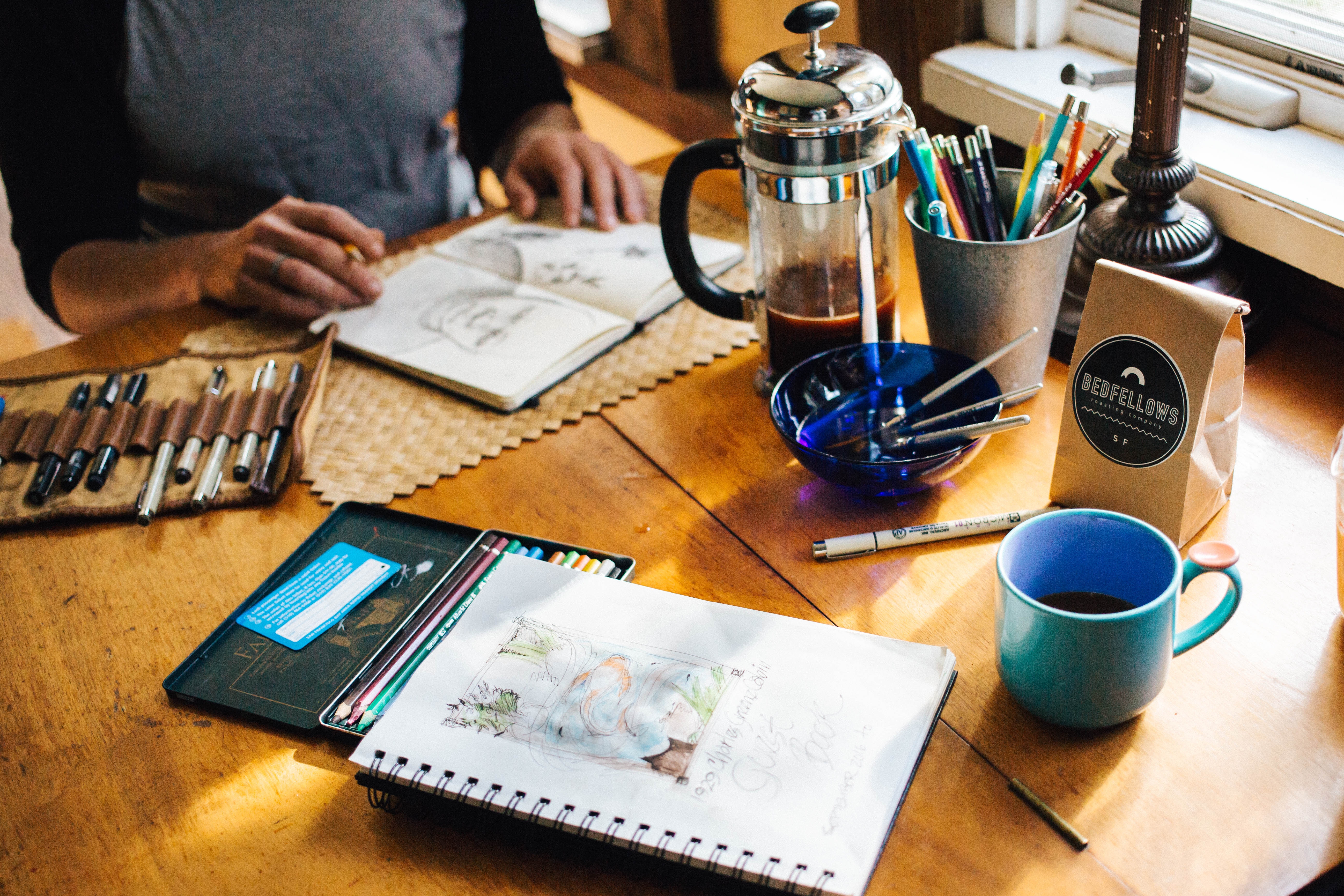 A person sketching at a table with coffee, colored pencils, a sketchbook, drawing supplies, and a French press.