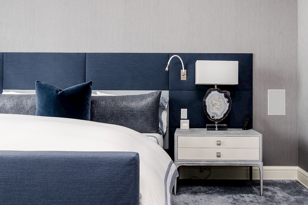 A modern bedroom features a blue headboard and bedding, a nightstand with a lamp, and decorative items on a grey carpet.