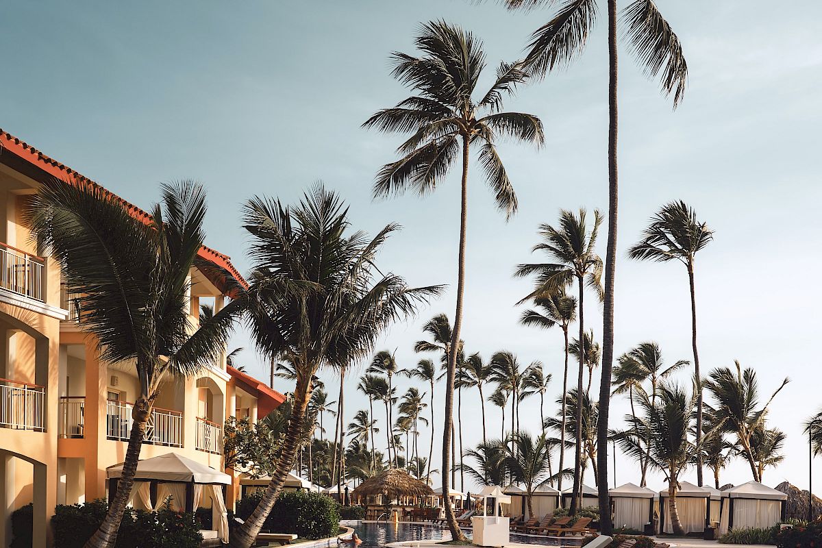 A serene resort pool area with tall palm trees, lounge chairs, and a building with balconies, all under a clear blue sky.